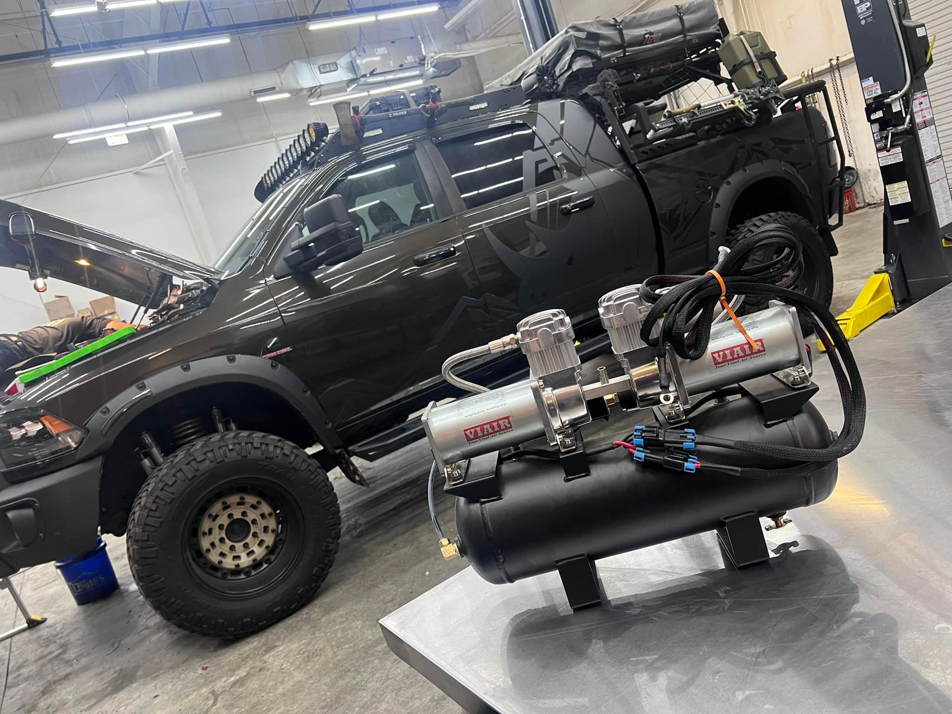 A black truck with the hood up is sitting in a garage next to a air compressor.