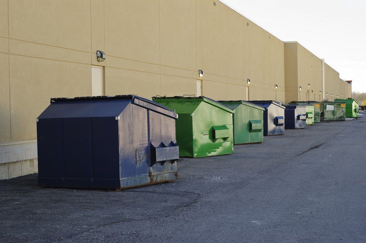 Dumpsters lined up against a beige building exterior. Various colors: blue, green, and gray.