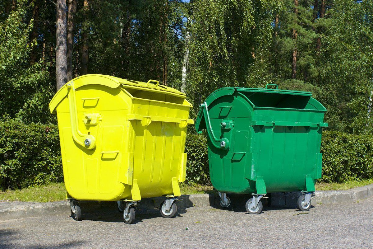 Yellow and green recycling bins on wheels on a paved surface, trees and greenery in the background.