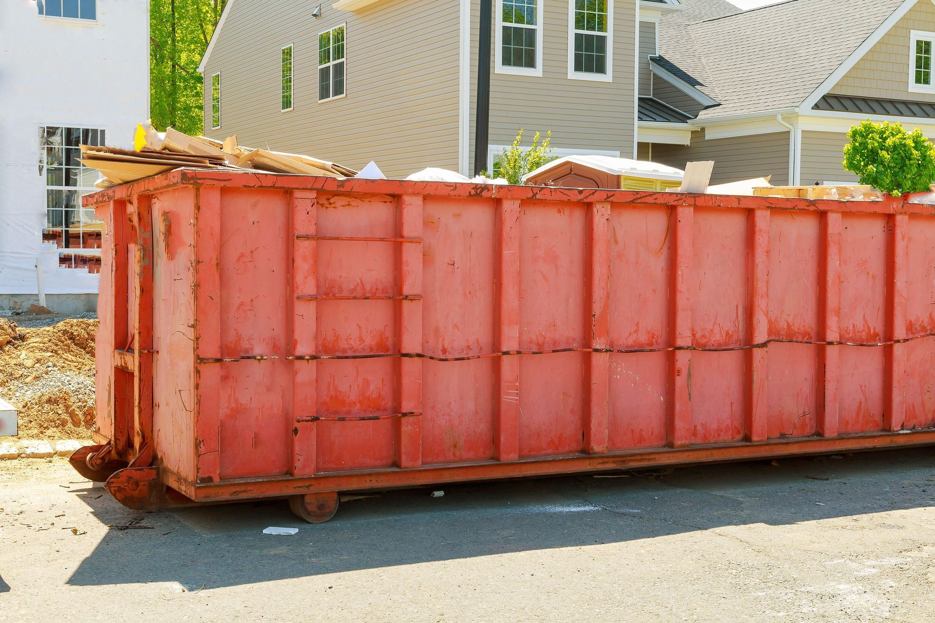 Red dumpster on a street, filled with construction debris, with houses under construction in the background.