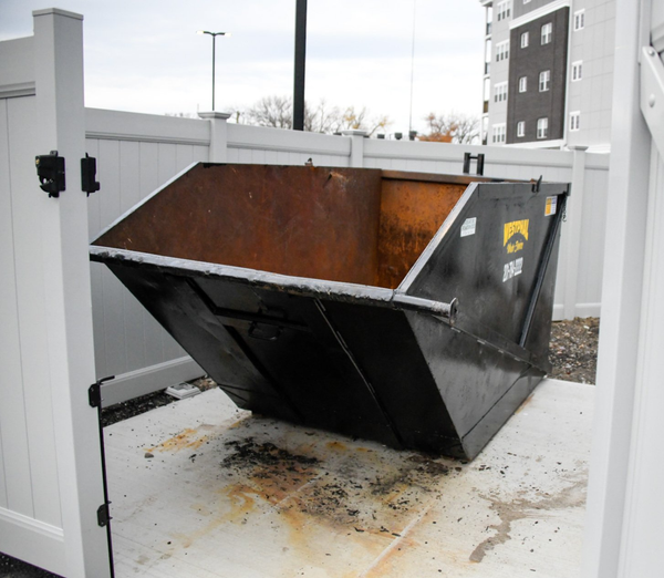 Black dumpster inside a white fenced enclosure on concrete.