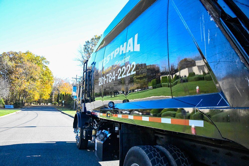 Black garbage truck on a road, with shiny blue side reflecting houses and trees.