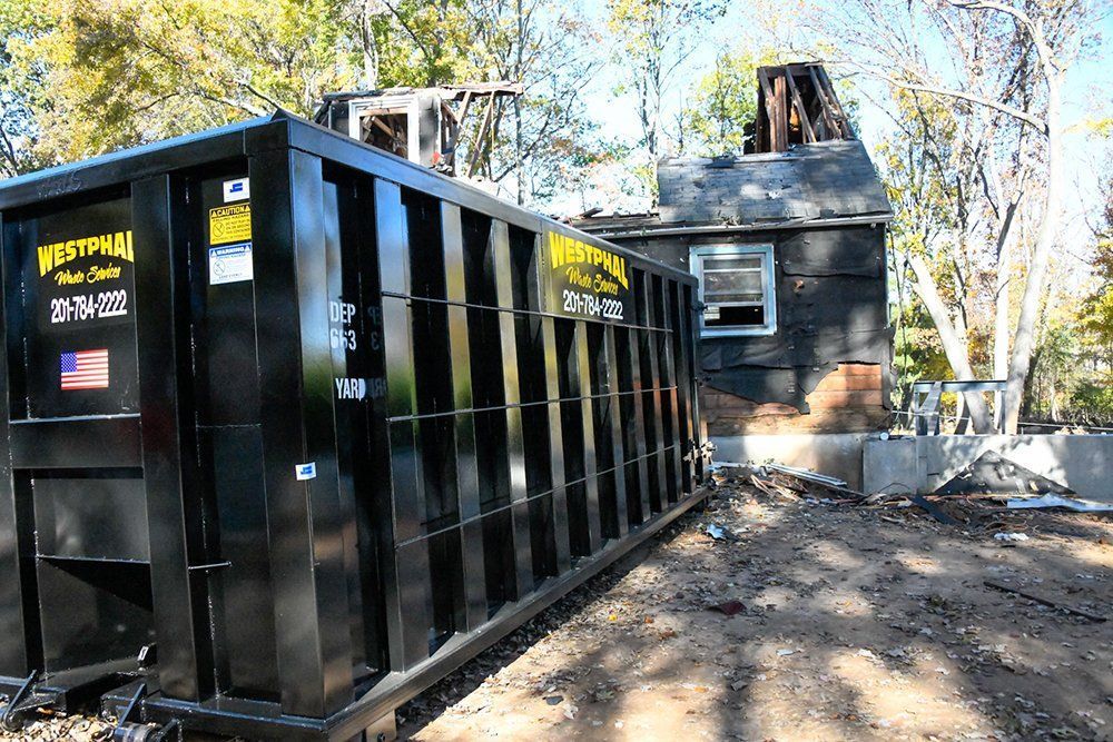 Black dumpster beside a partially burned house on a sunny day.