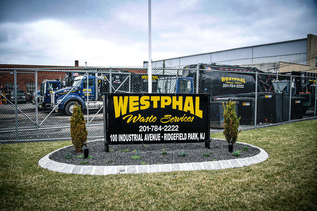 Sign for Westphal Waste Services, address, and phone number, with trucks behind a fence.