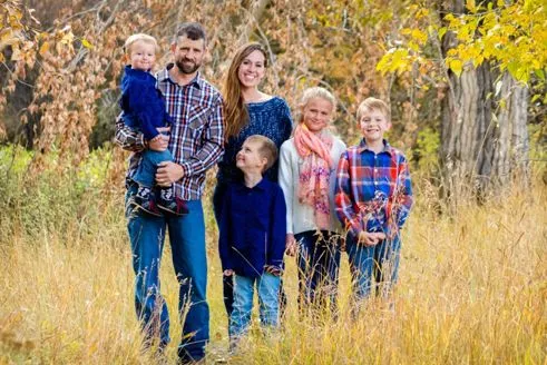 Family of six in field, autumn foliage backdrop. Adults and children smiling, wearing casual clothes.