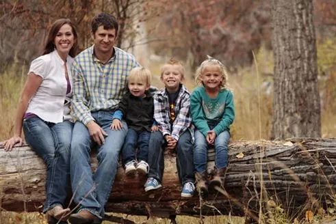 Family of five smiling, sitting on a log outdoors in autumn.