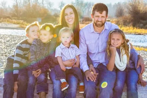 Family of seven smiling outdoors, sitting on log. River in the background. Sunny.