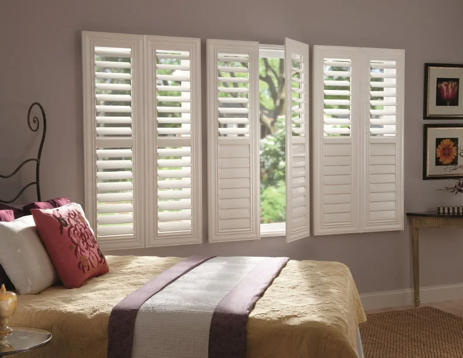 Bedroom with white shutters, partially open, letting in light. Bed with pillows and blanket in foreground.