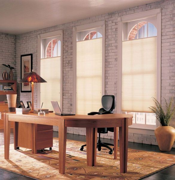Office interior with three arched windows, light tan shades, wooden desk and furniture, brick walls.