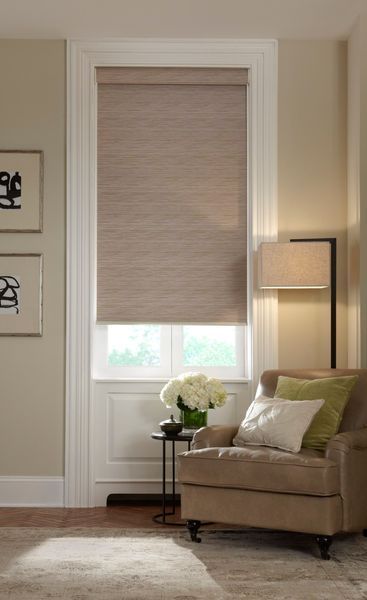 Beige roller shade in a window, over a small table with a floral arrangement and a beige armchair.