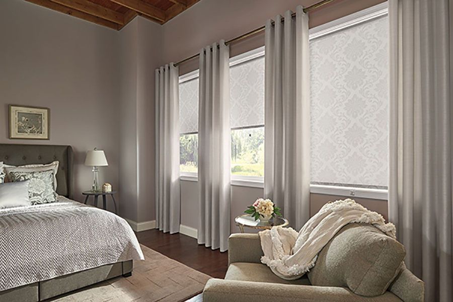 Bedroom with bed, armchair, windows with patterned shades and curtains, neutral color scheme.