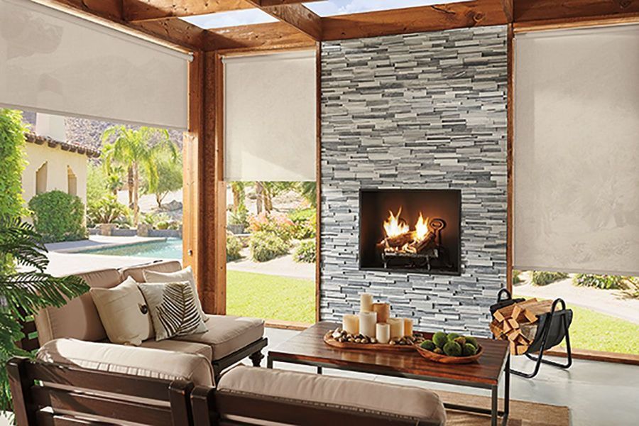 Cozy outdoor living space with fireplace, stone wall, and light-colored shades. Furnished with a sofa and coffee table.