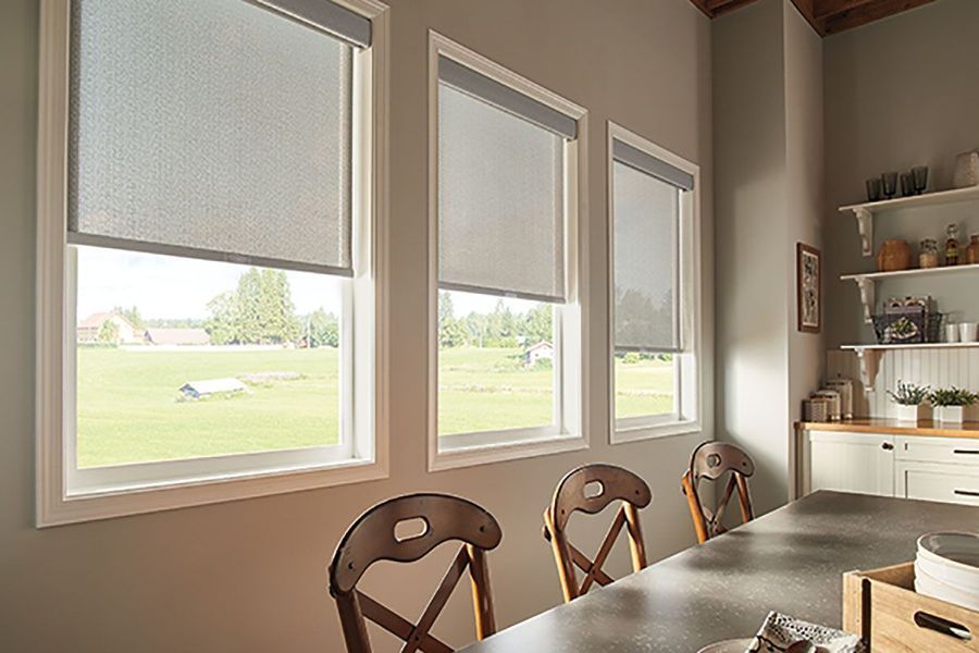 Three windows with gray roller shades in a kitchen, overlooking a green field. Dining table and chairs in foreground.