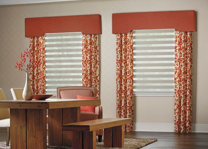 Dining room with two windows. Orange valances, patterned drapes, and horizontal blinds. Wooden table and bench.