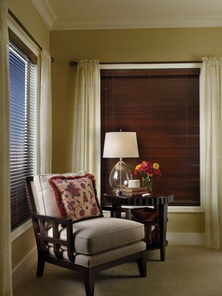 Cozy corner with a wooden armchair, end table with lamp, flower bouquet, and two windows with blinds and curtains.