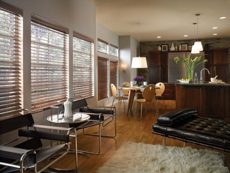 Living room with wooden blinds, hardwood floor, modern furniture, and kitchen visible in the background.