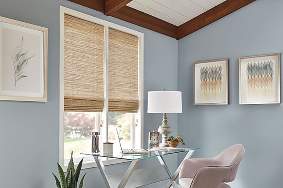 Home office with blue walls, window with woven shade, glass desk, artwork, and pink chair.