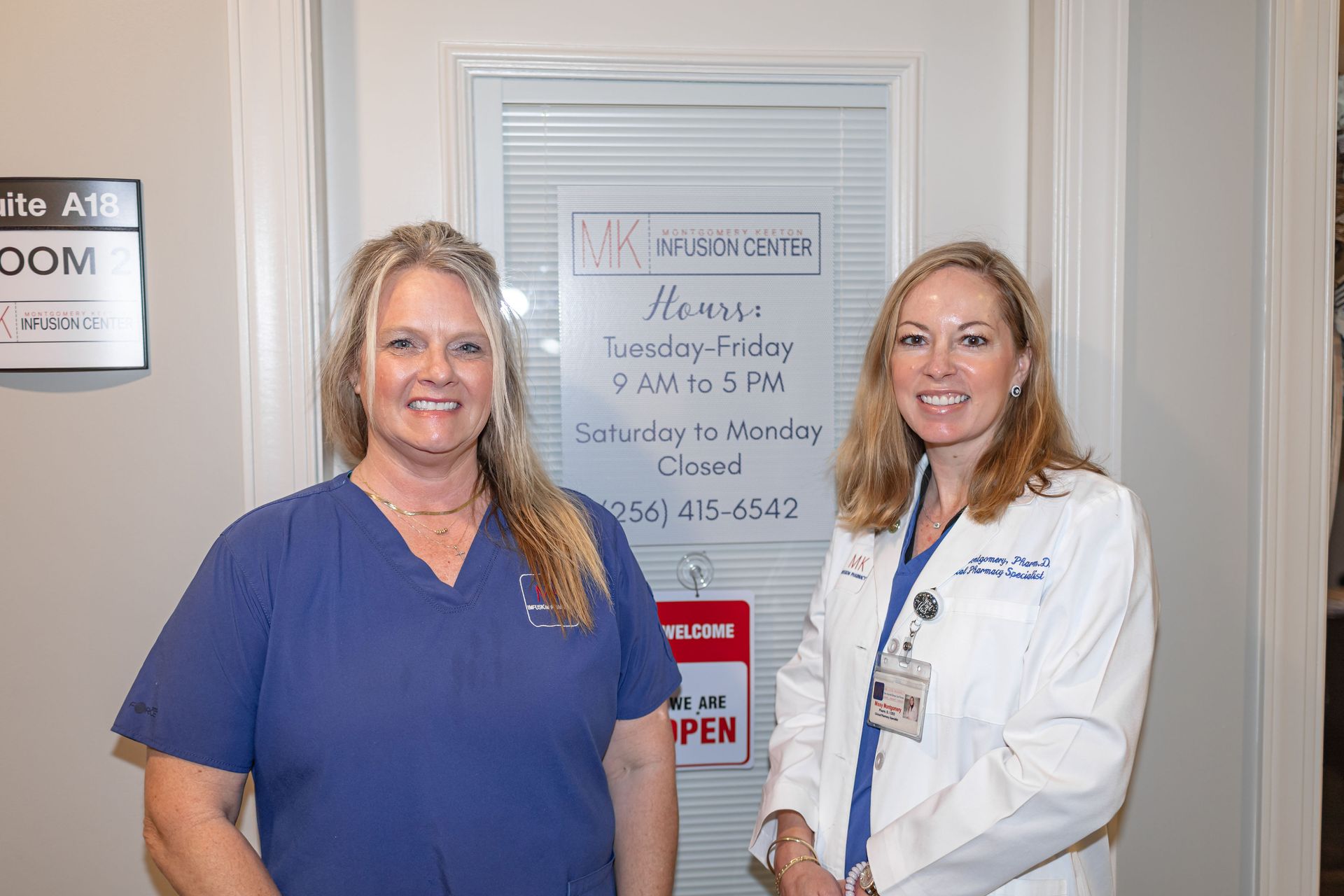 Two women stand in a clinic hallway, one in blue scrubs and one in a white coat.