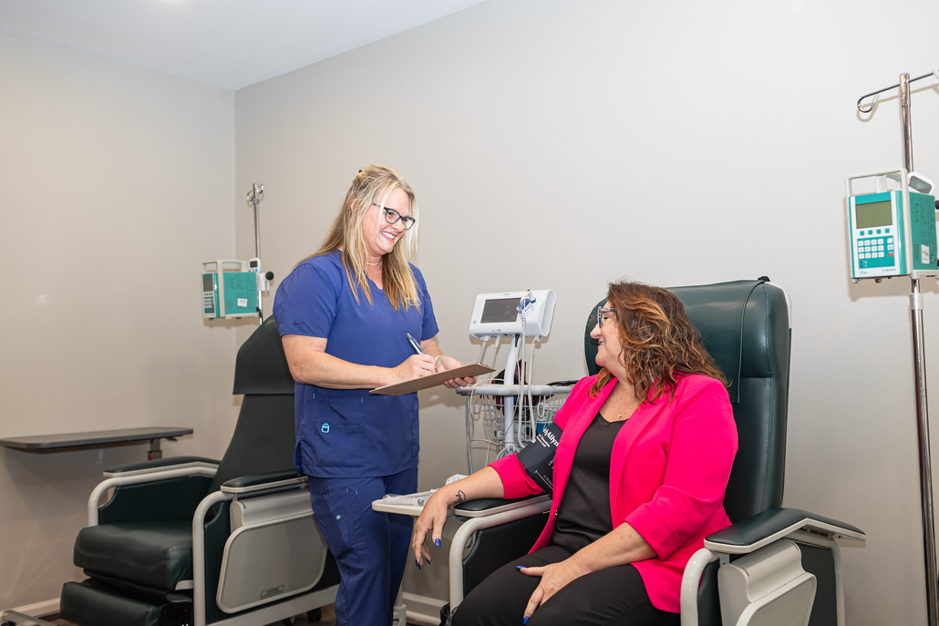 Nurse in blue scrubs speaking with a patient seated in a clinic chair during an infusion treatment.