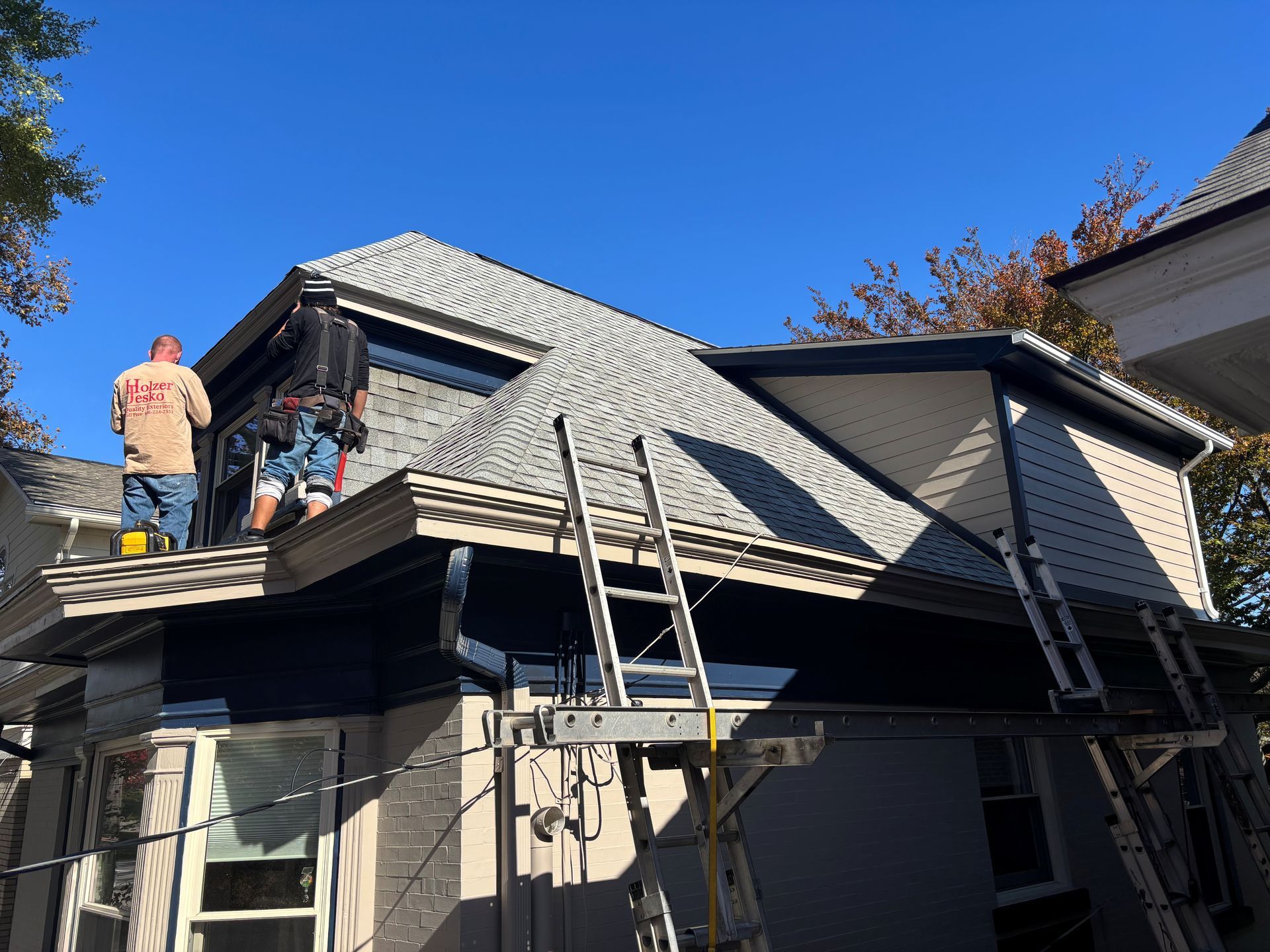 Two workers on a roof installing shingles, ladders, blue sky.