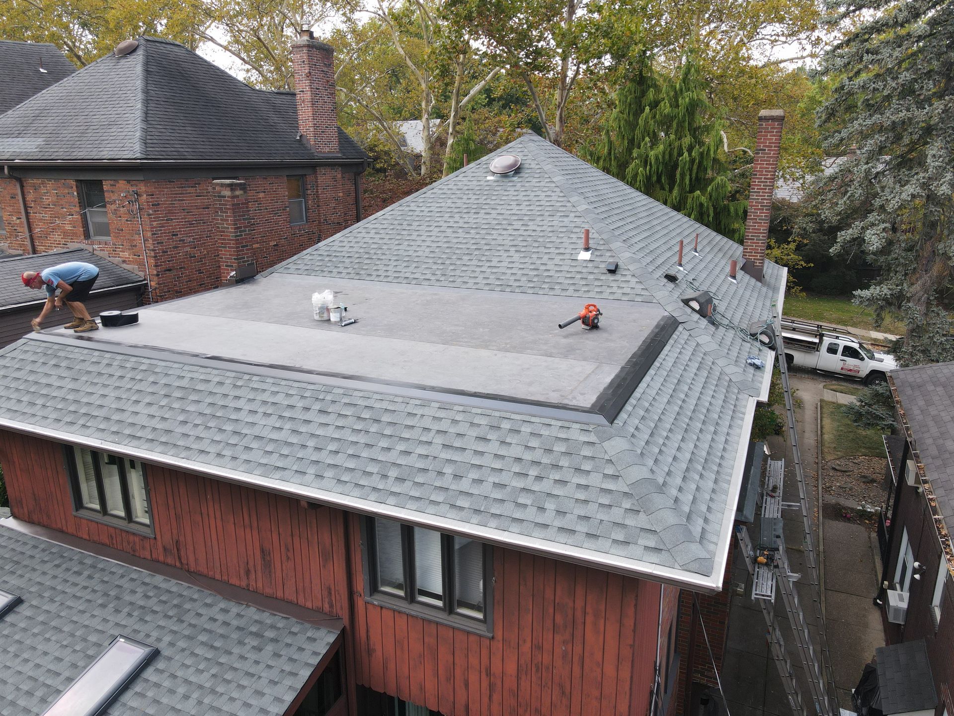 A house with a gray shingle roof. A worker is installing shingles. A truck is parked nearby.