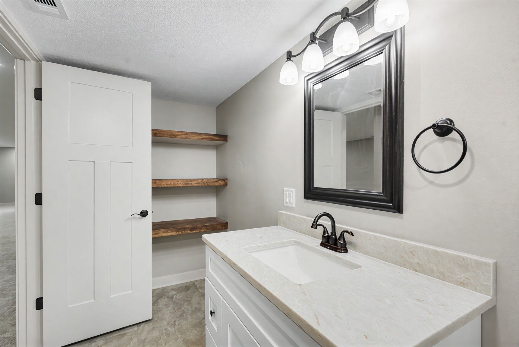 Bright bathroom with white vanity, dark mirror, towel ring, and open shelves by a closed door.