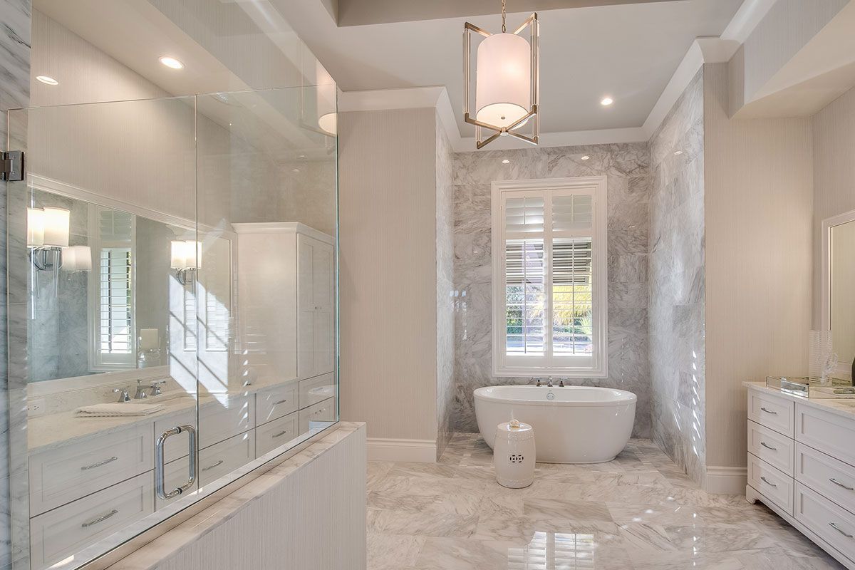 Bright luxury bathroom with marble floors, glass shower, double vanities, and a freestanding tub by a window.