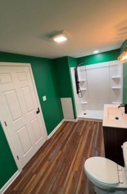 Bright green bathroom with white vanity, shower, toilet, and wood-look floor.