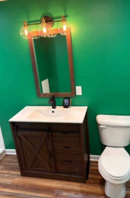 Bathroom vanity with white sink, wood cabinet, mirror, and toilet against a green wall