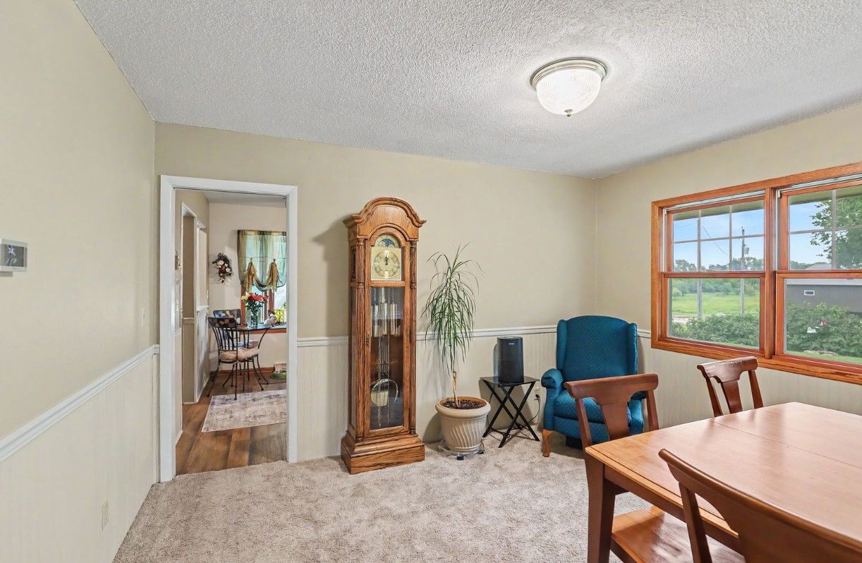 Bright living room with carpet, grandfather clock, blue chair, dining table, and large window.
