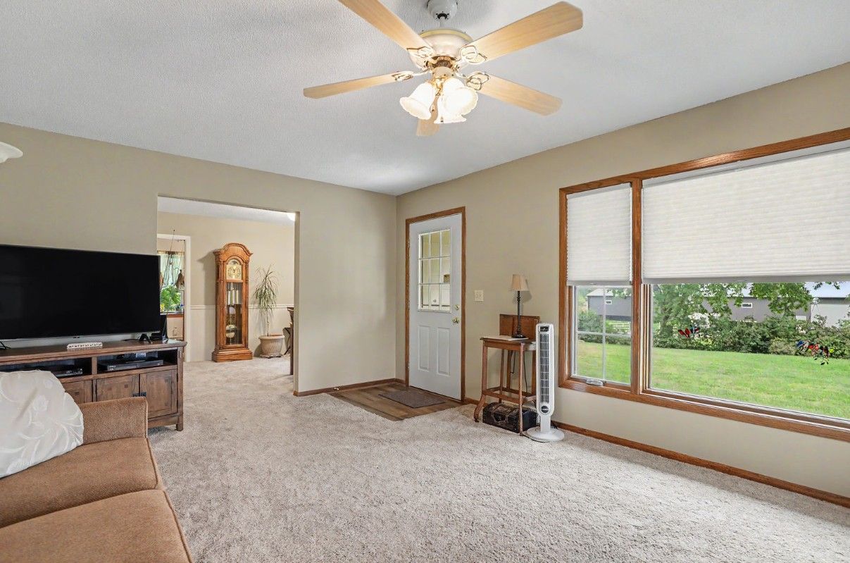 Carpeted living room with TV, ceiling fan, and large window overlooking a green yard.