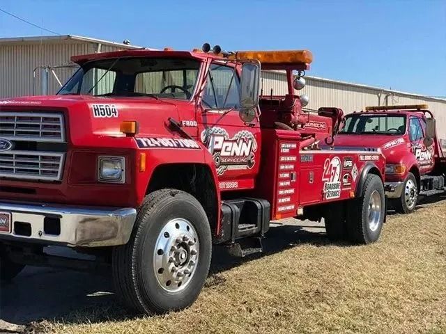 Red tow truck with attached flatbed trailer, towing a red pickup truck outdoors on a sunny day.