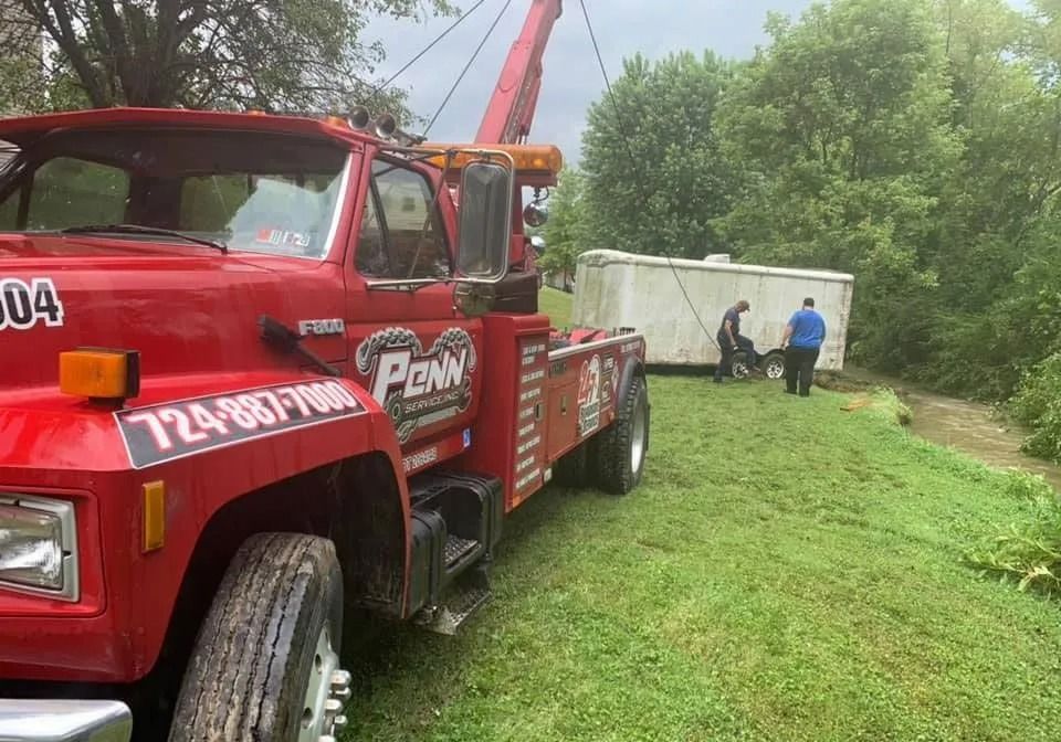 Red tow truck towing a white container next to a grassy hill. Two people stand near the container.