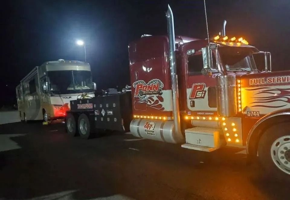 A tow truck pulling a beige RV at night. The truck is red and has bright lights.