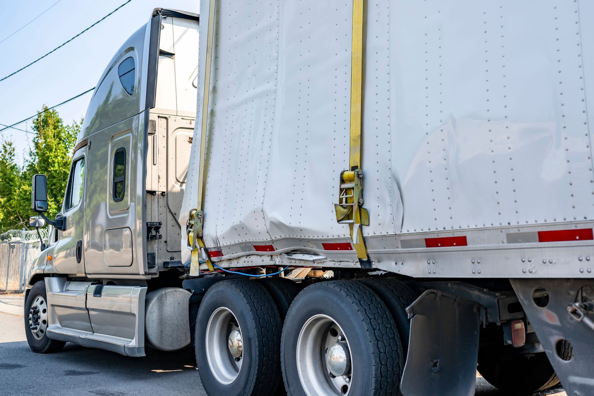 Semi-truck with a damaged trailer on a road. Gray cab, white trailer, black tires.