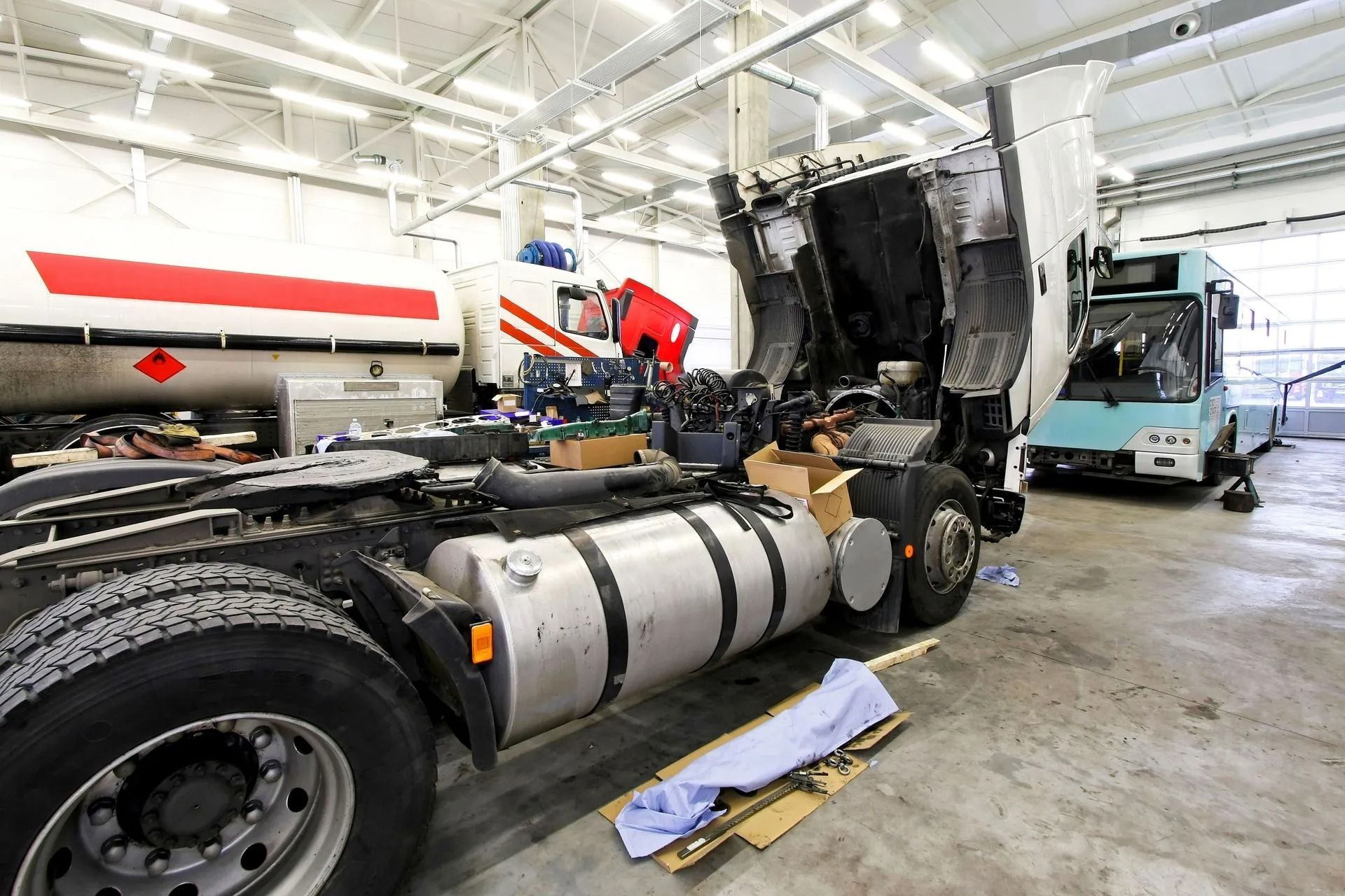 Truck in a repair shop with hood open, another truck and a bus visible.