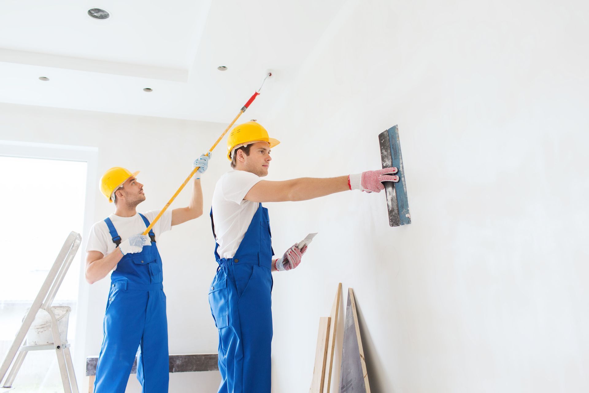 Two workers in blue overalls and yellow hard hats plastering a white wall in a room.
