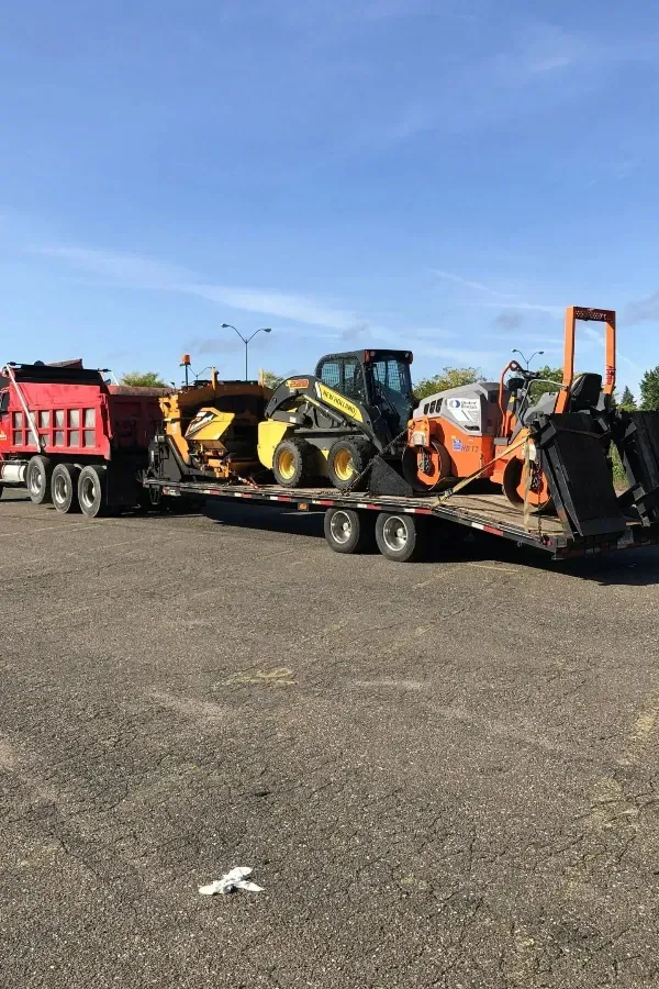 A truck is carrying a dumpster and a tractor on a trailer