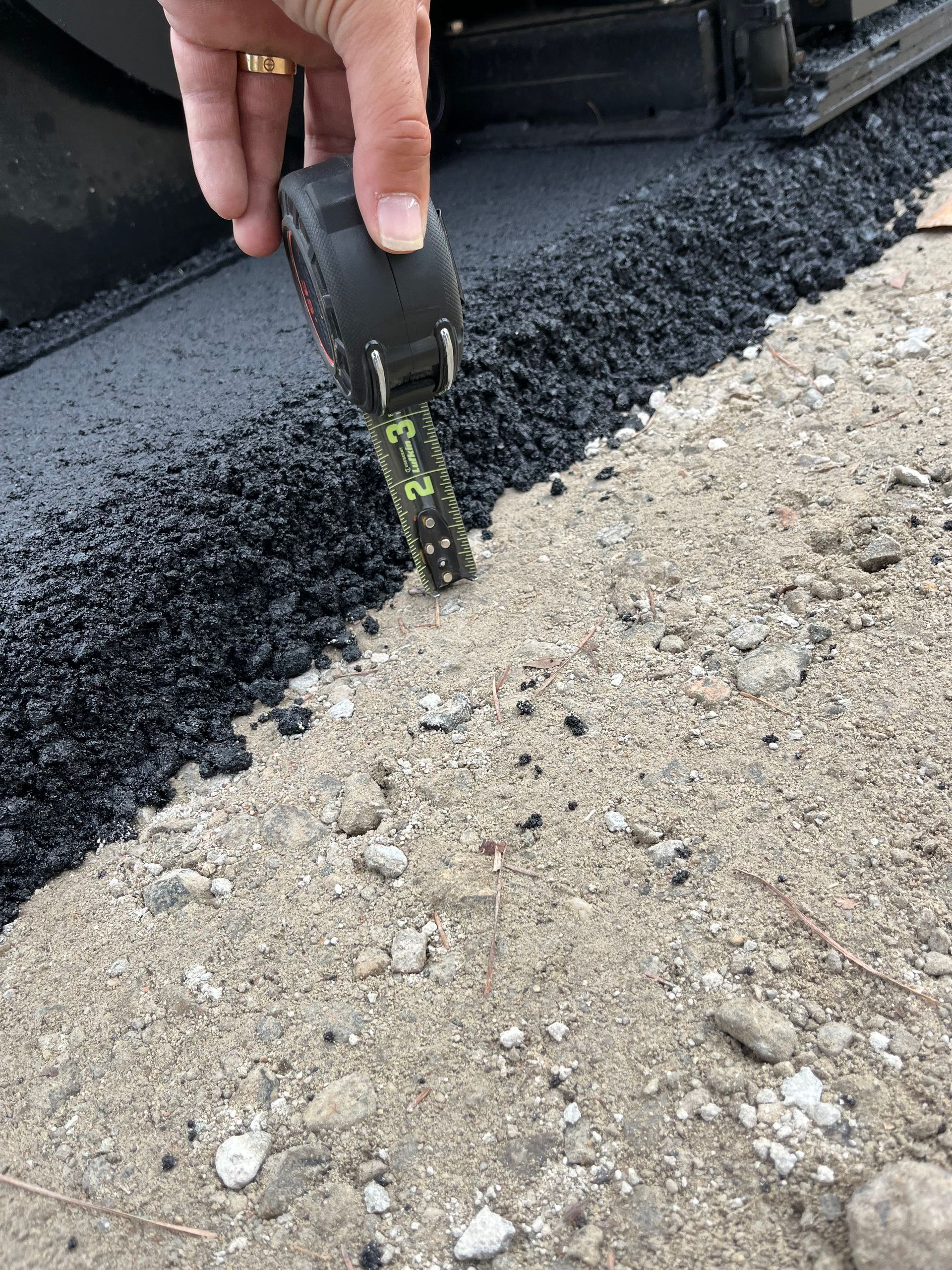 A person is measuring the depth of asphalt with a tape measure
