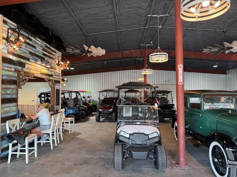 Open-air garage with golf carts, a seated person, and a vintage green car under a black ceiling