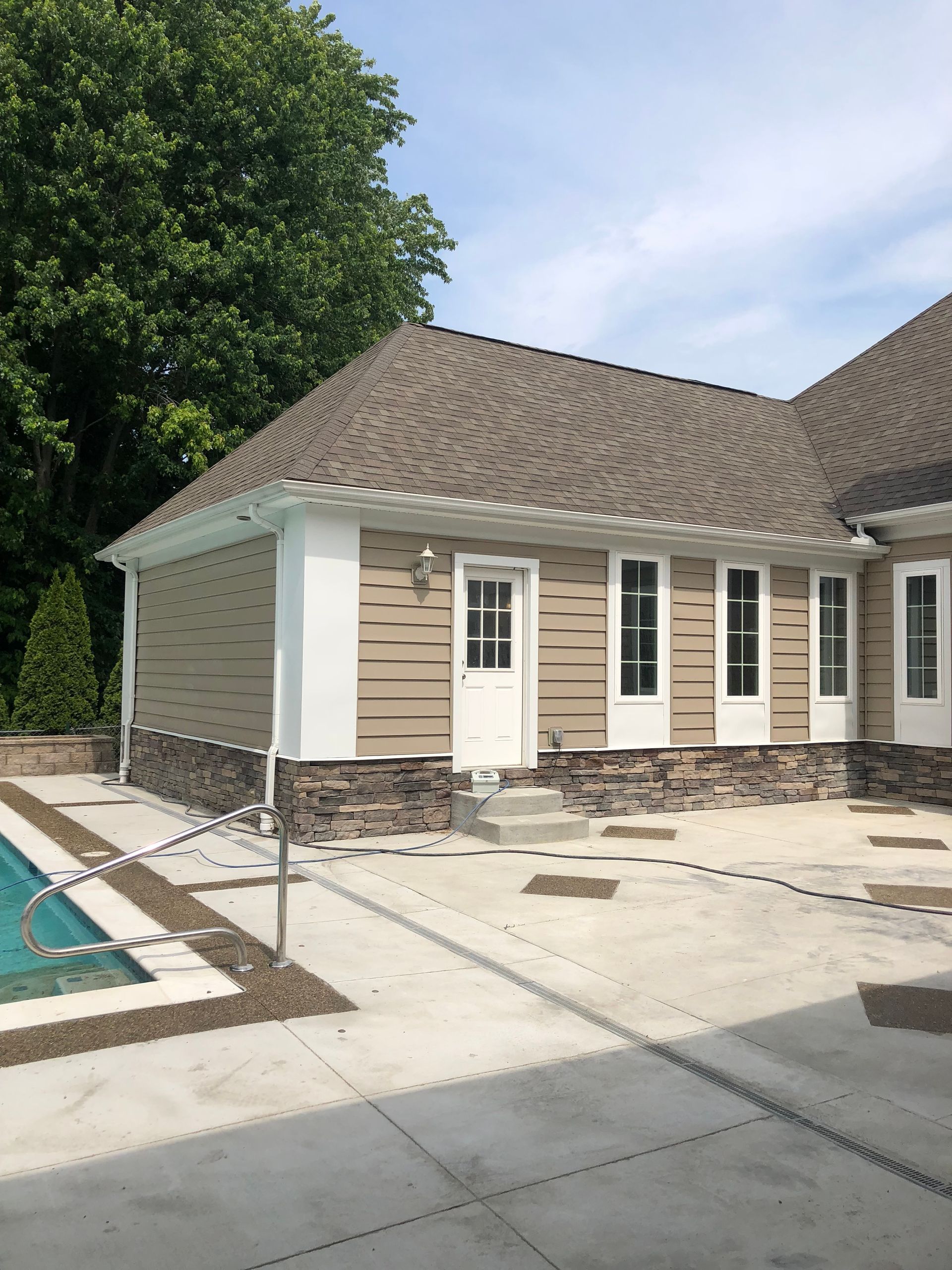 Backyard pool beside beige house with stone trim and white patio in bright daylight