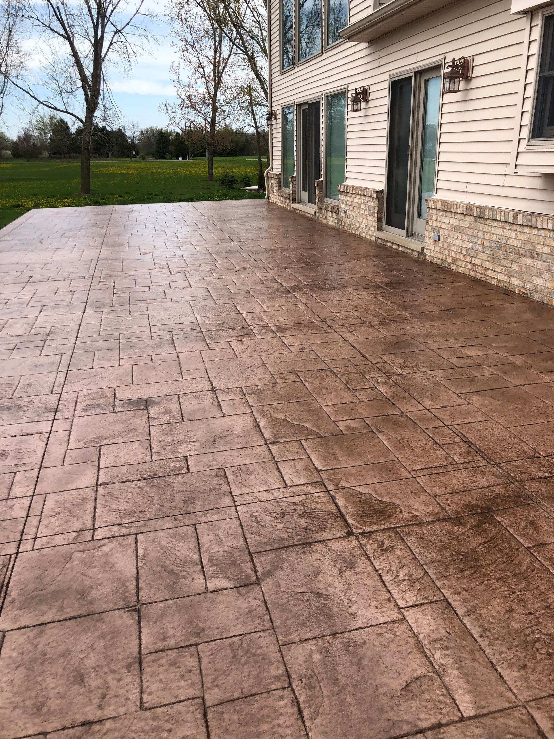Stamped concrete patio beside a beige house with teal shutters, overlooking a grassy yard and trees