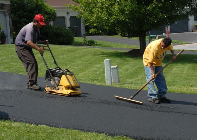 Two workers paving a driveway, one using a roller and one smoothing fresh asphalt