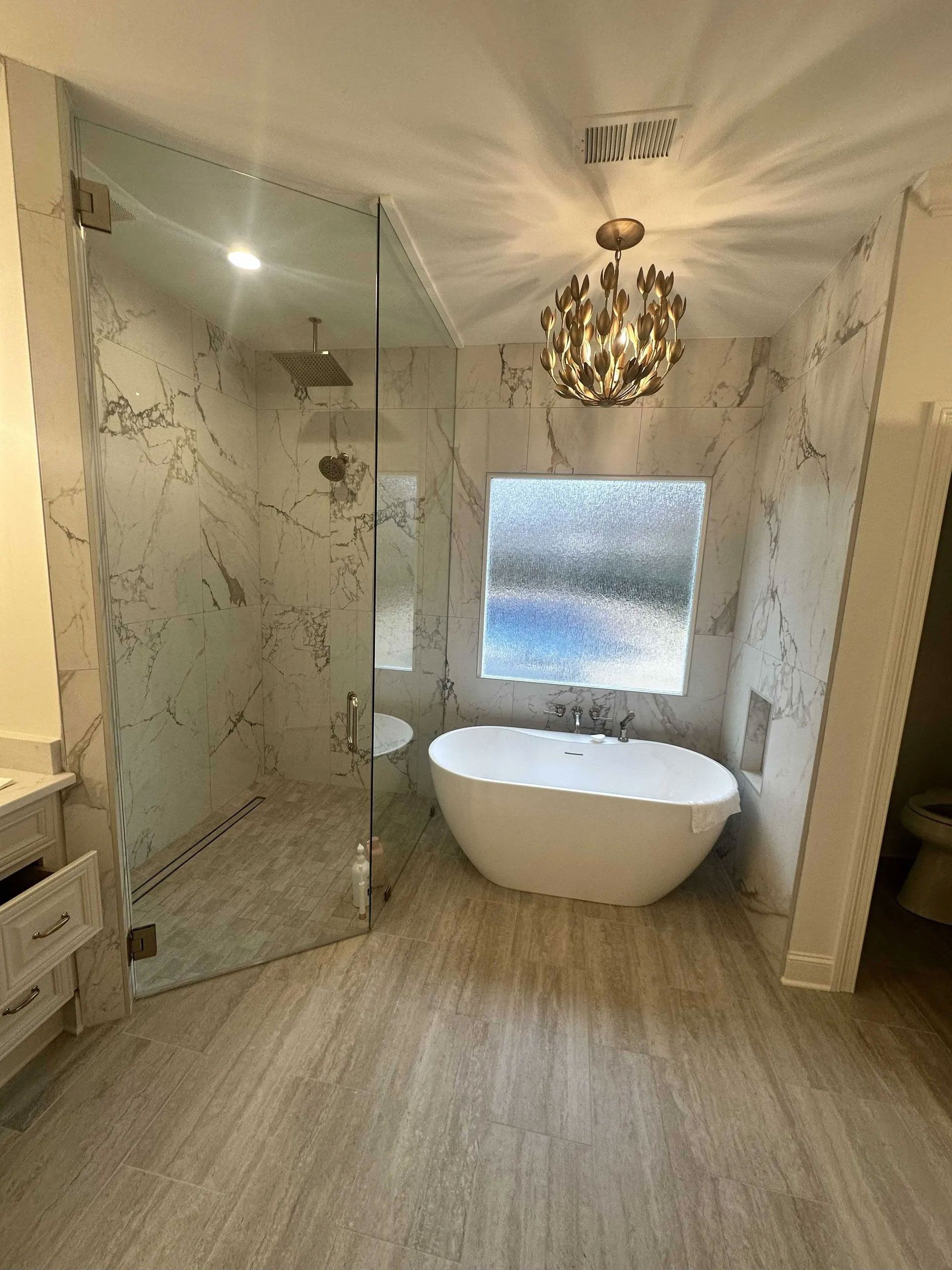 A modern bathroom featuring a white freestanding tub, a large glass-enclosed shower, marble-patterned walls, and a chandelier.