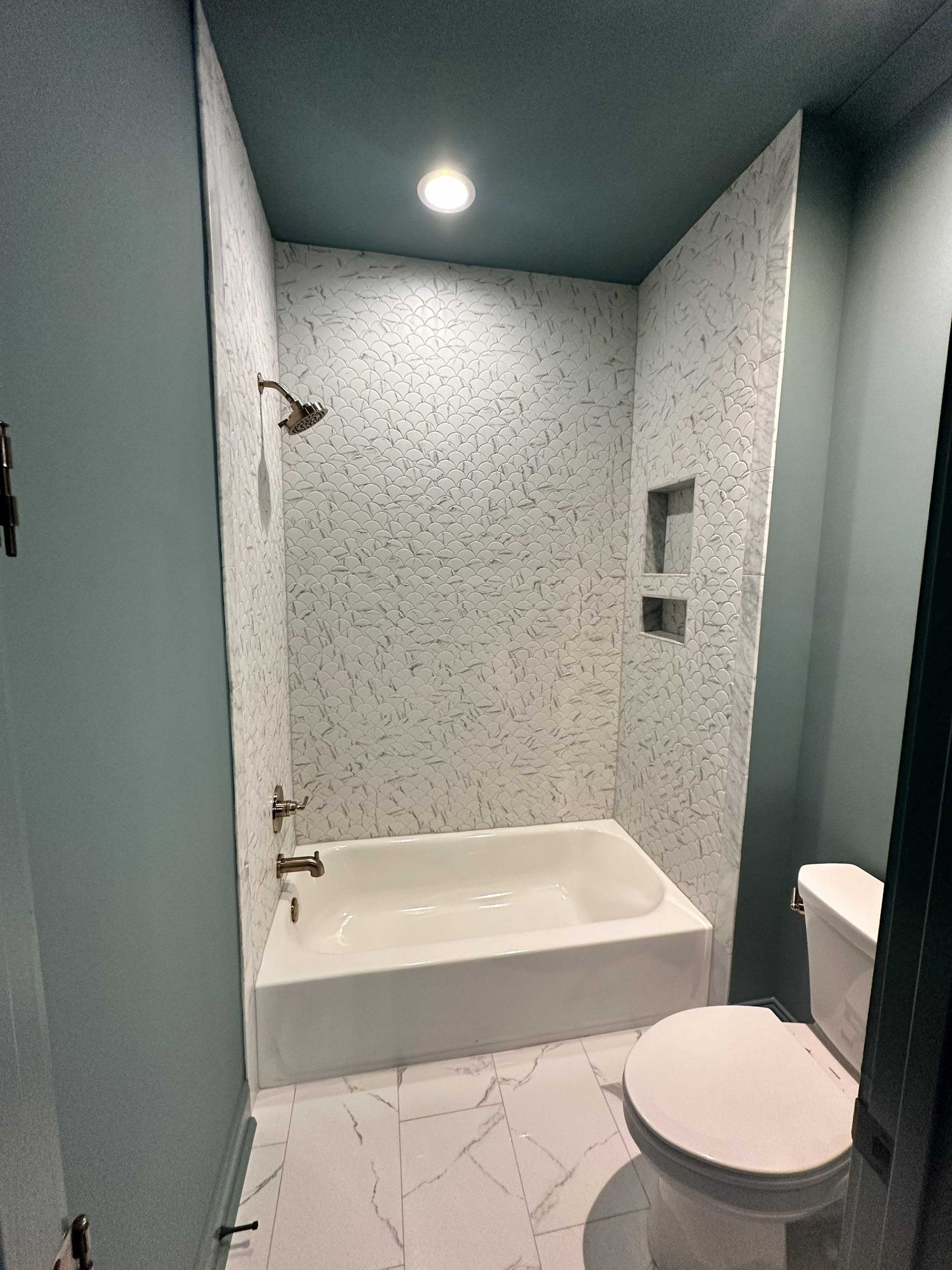 A small bathroom with a white tub, mosaic shower walls, a teal ceiling, and a toilet.