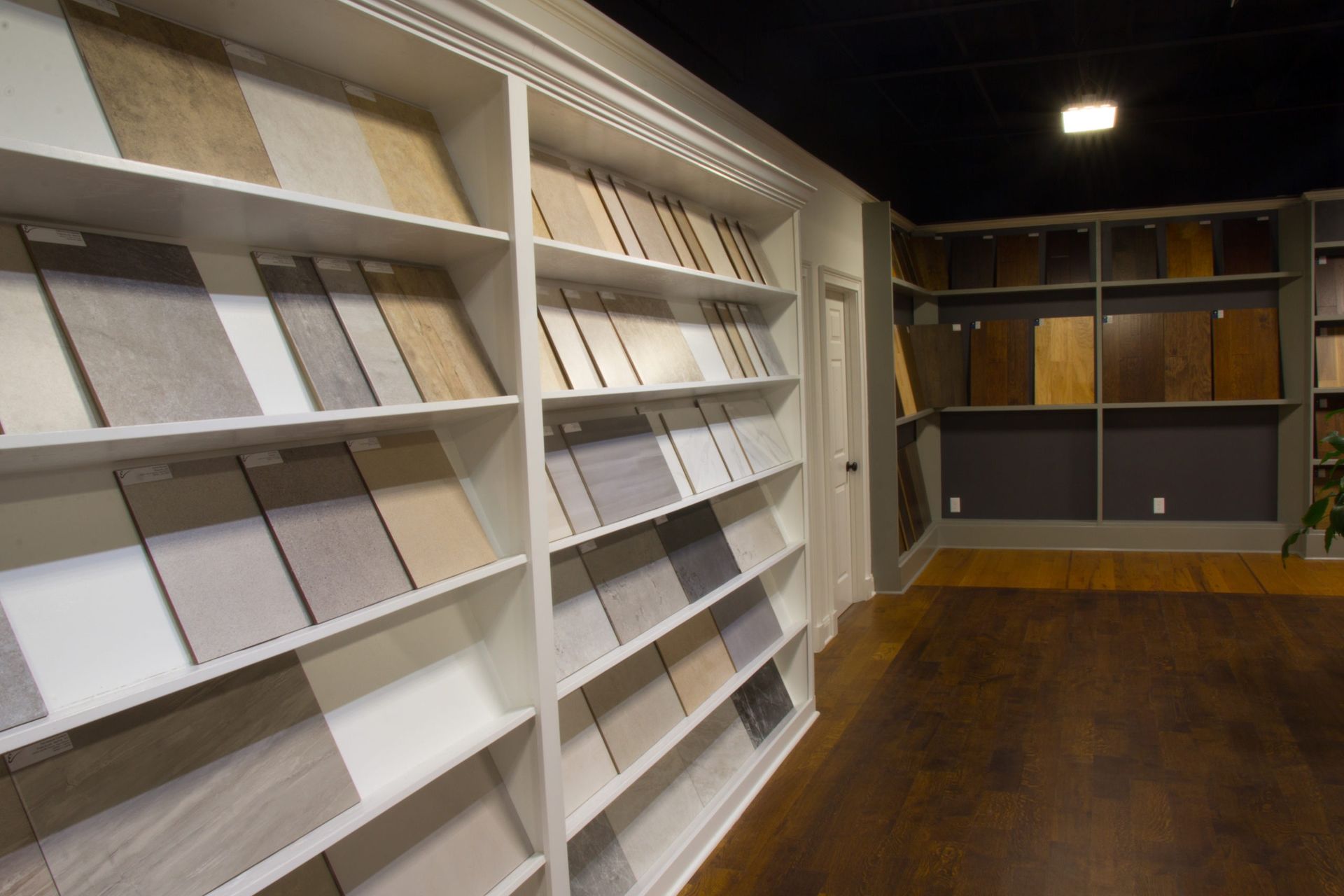 Tile samples displayed on white shelves in a showroom; dark wood flooring.