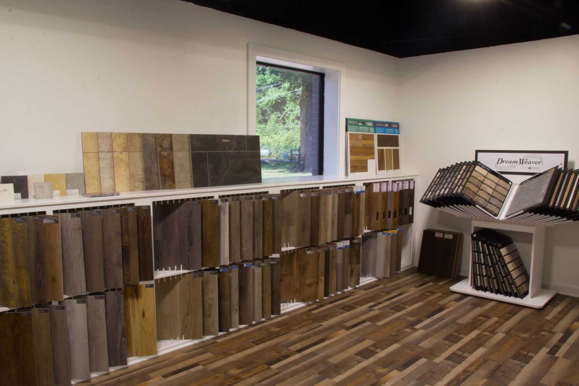 Flooring samples displayed in a showroom with a window, showcasing various colors and textures of wood and tile.
