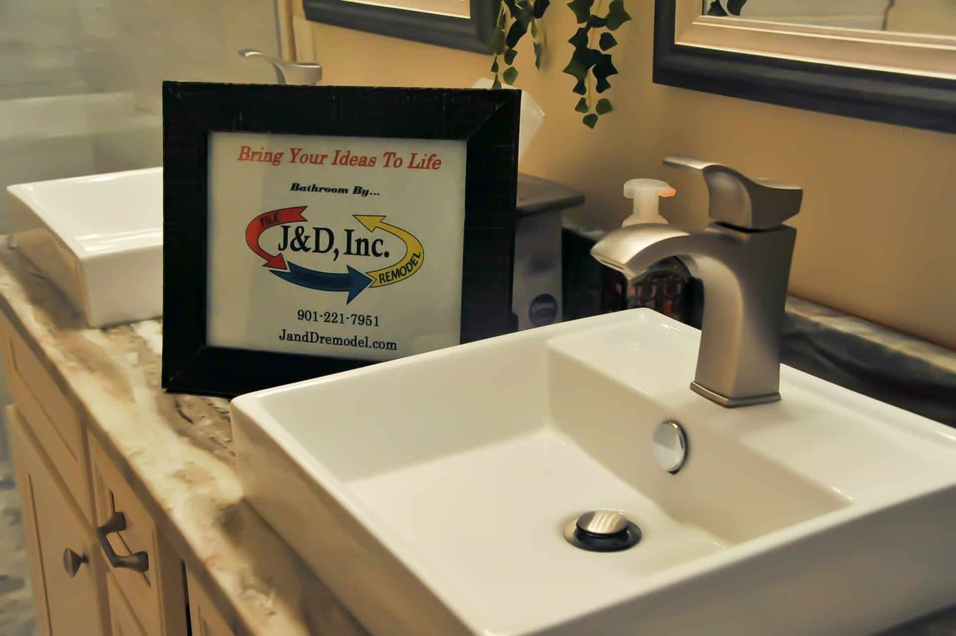 Bathroom vanity with white square sink, faucet, framed company logo, and beige countertop.
