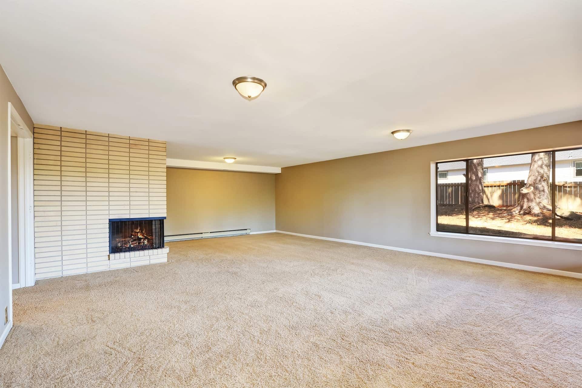 Empty living room with tan carpet, a brick fireplace, and a large window.