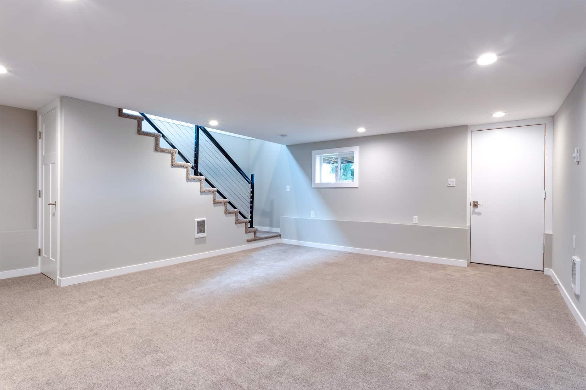 Empty basement with gray walls, beige carpet, stairs, a window, and a white door.